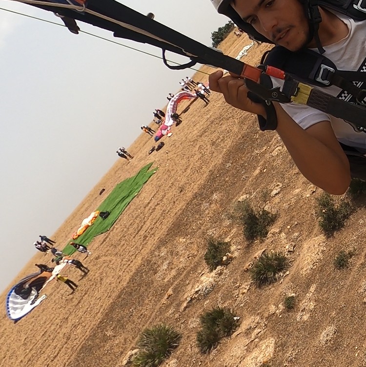 A person holding a paraglider in a grassy, outdoor landscape with others in the background.
