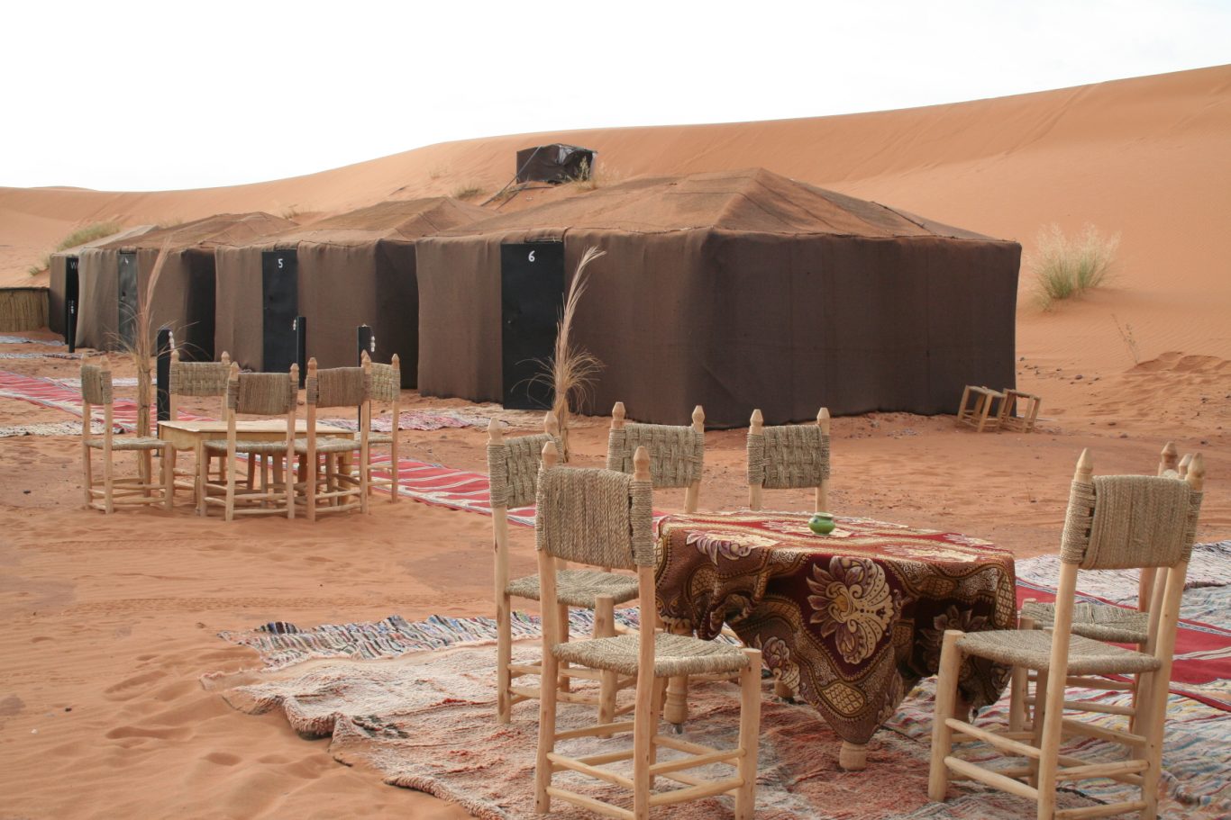 Merzouga camp Tents set up on sand dunes with wooden tables and chairs arranged for dining.